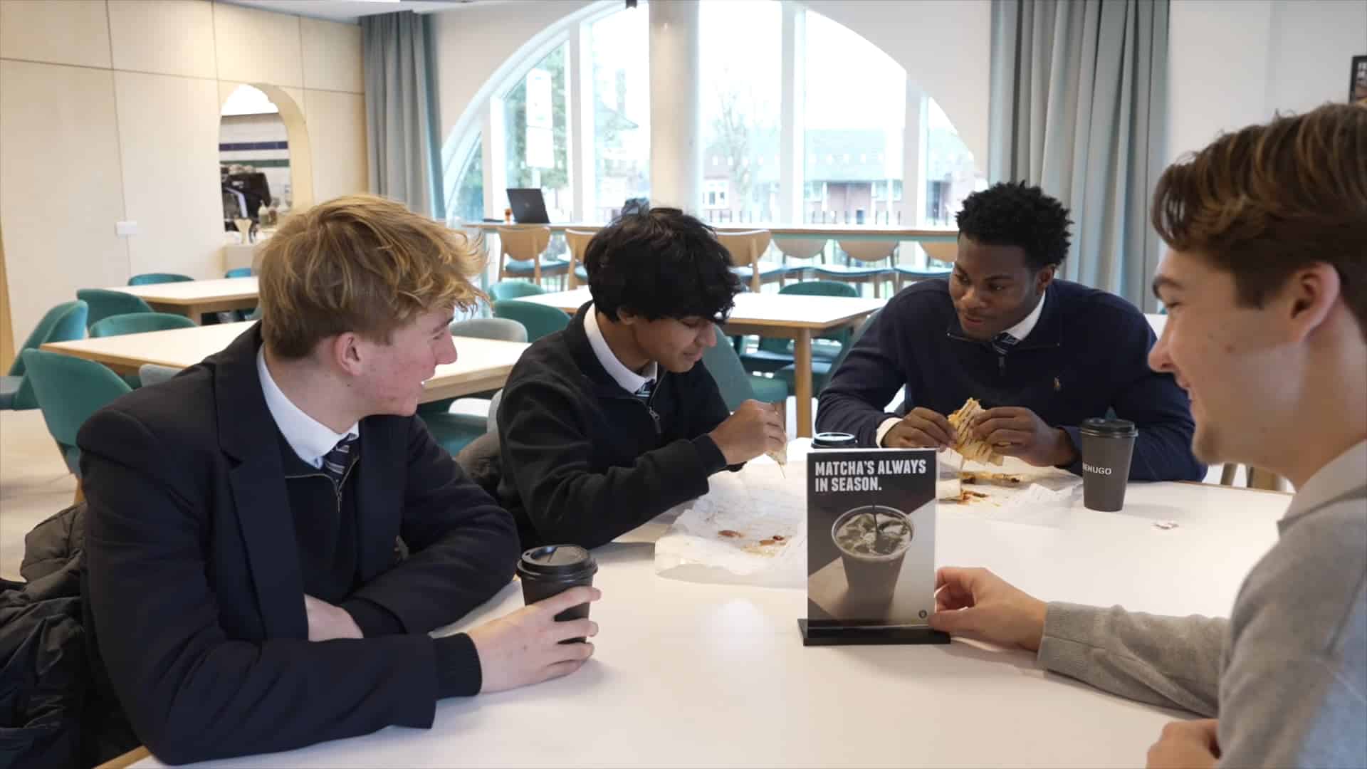Four students in school uniforms sit around a table in the New Boarding House canteen, eating and chatting. Three have food and drinks, while one holds a coffee cup. Large windows and teal chairs are visible in the background.