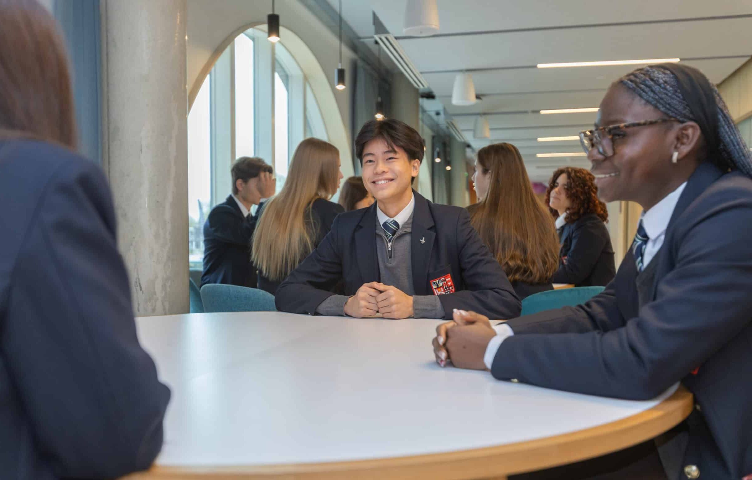 A group of students in uniforms sit around a round table in a bright, modern classroom at the New Boarding House, smiling and talking with each other.