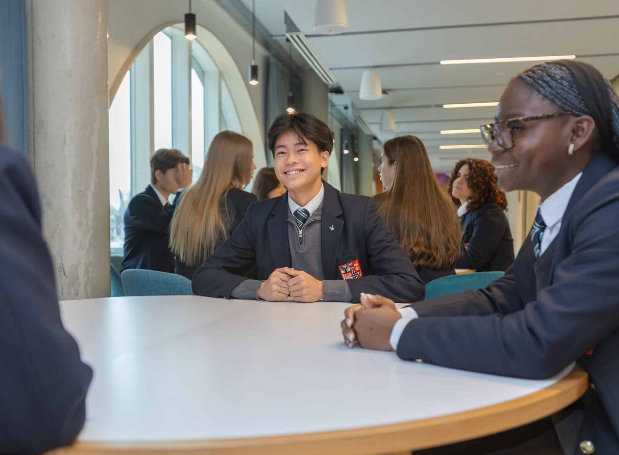 A group of students in uniforms sit around a round table in a bright, modern classroom at the New Boarding House, smiling and talking with each other.