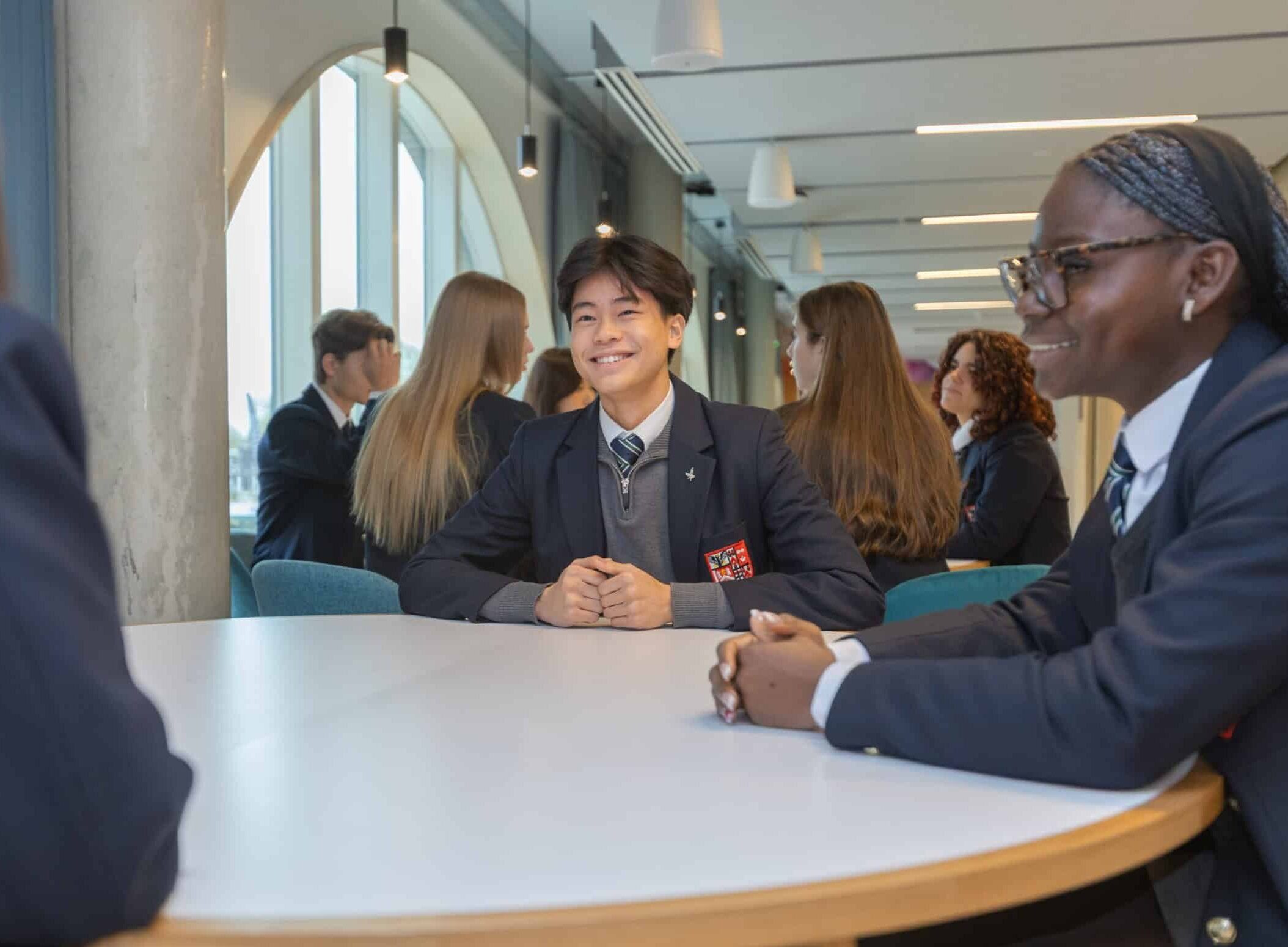 A group of students in uniforms sit around a round table in a bright, modern classroom at the New Boarding House, smiling and talking with each other.