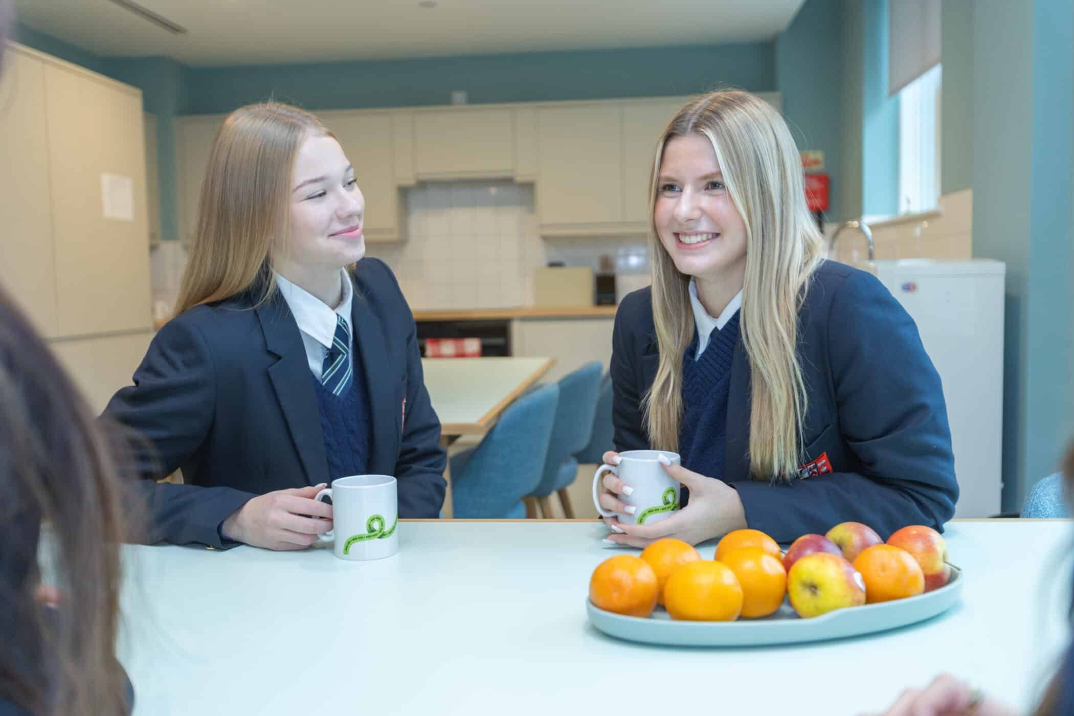 Two teenage girls in school uniforms sit at a table, smiling and holding mugs. A plate of oranges and apples is on the table in front of them. They appear to be in a bright, modern kitchen or staff room.