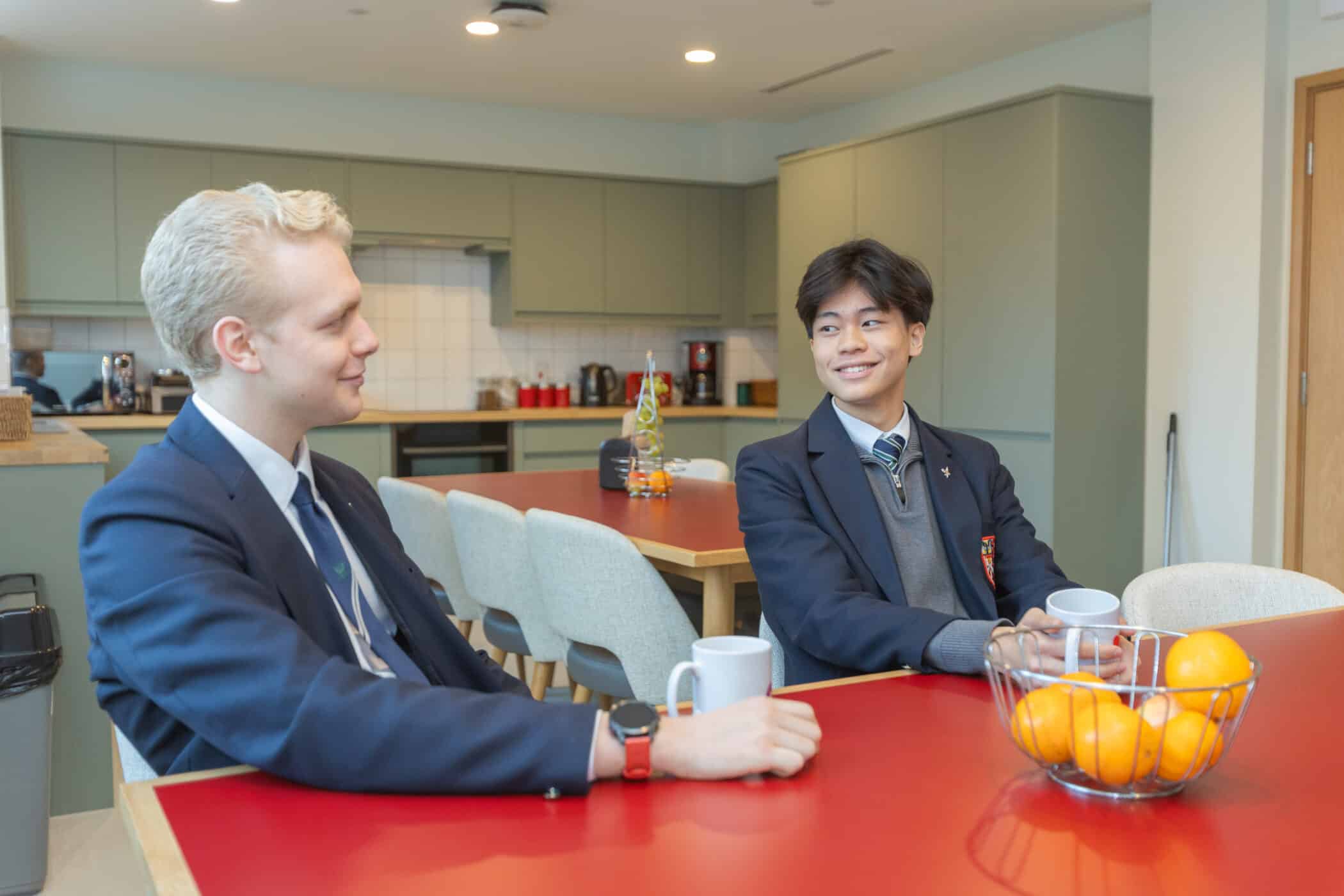 Two young men in suits sit at a red table in a modern kitchen, smiling and talking over white mugs. A bowl of oranges sits between them, a friendly scene reflecting the camaraderie found whilst boarding at Brentwood.