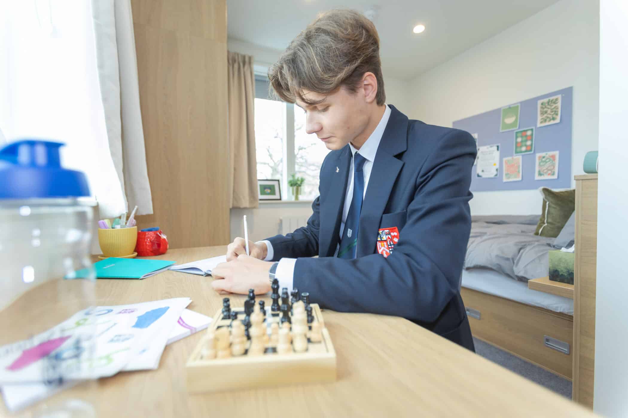 A young student in a school uniform sits at a desk in a bedroom, writing in an exercise book. A chess set, papers, and stationery are on the desk, and the bed and a noticeboard are visible in the background.
