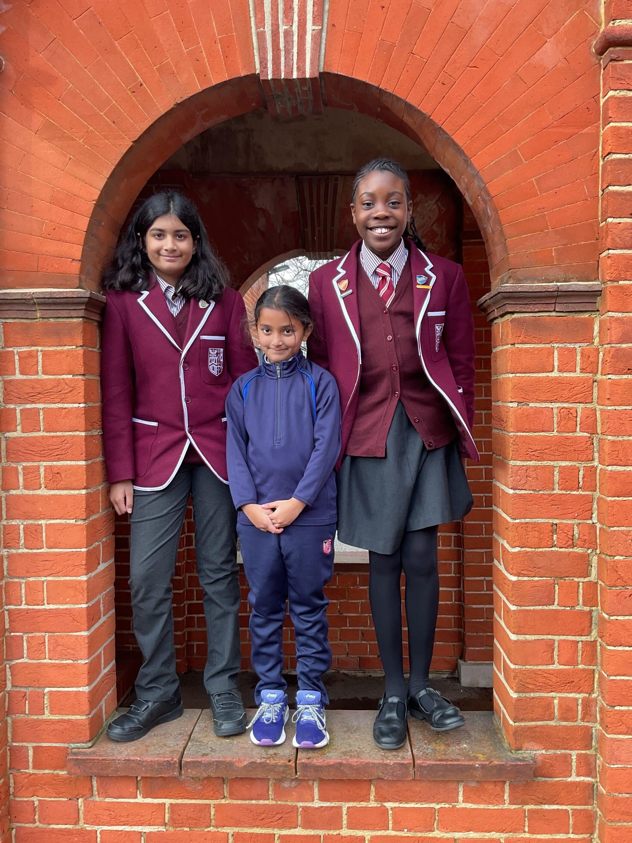 Three children pose and smile under a brick archway. Two older girls wear matching maroon school blazers and grey skirts or trousers, and a younger child in the middle wears a blue tracksuit and trainers.