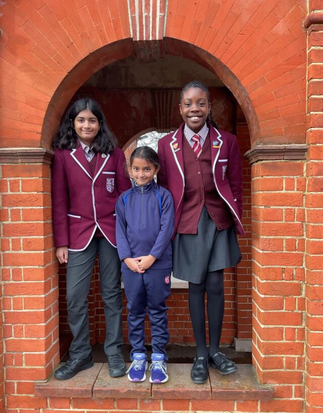 Three children pose and smile under a brick archway. Two older girls wear matching maroon school blazers and grey skirts or trousers, and a younger child in the middle wears a blue tracksuit and trainers.