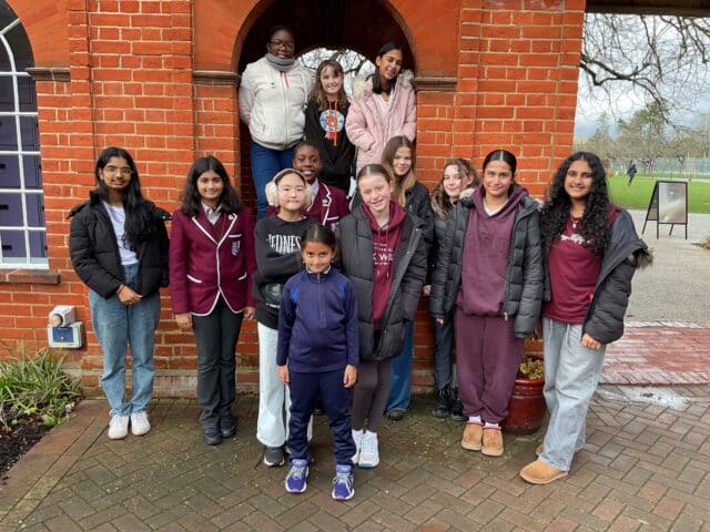 A group of children pose outside in front of a brick building with an arched doorway. Most wear jackets or school uniforms and smile at the camera. Trees and a park can be seen in the background.