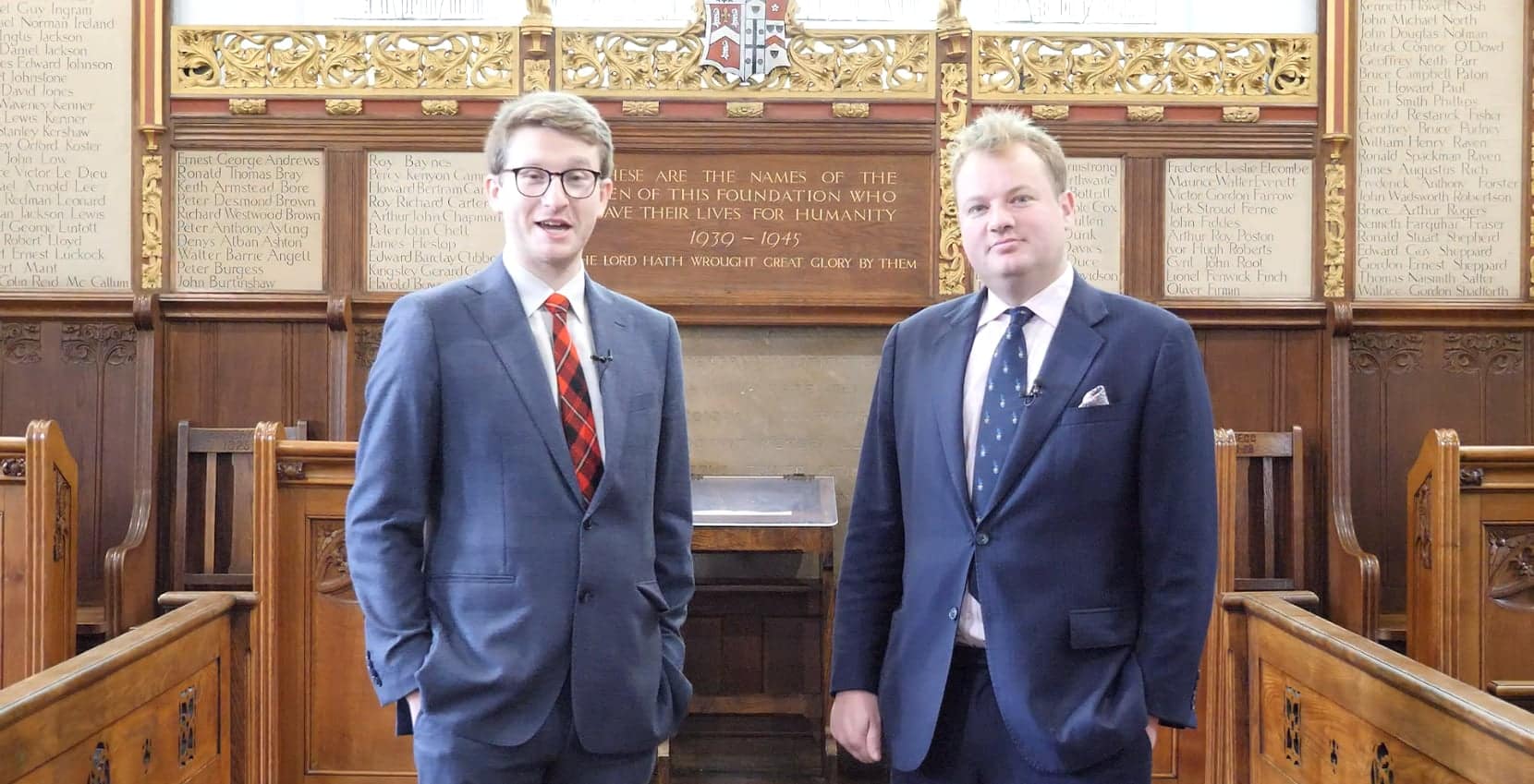 Two men in suits stand inside a wooden-panelled hall with memorial plaques behind them; the setting appears formal and historical, suggesting an event like the Brentwood International Music Competition.