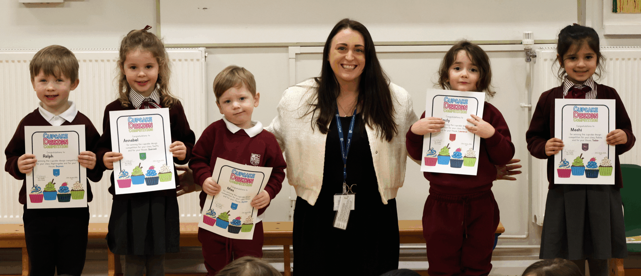A woman stands smiling with five young children in school uniforms. The children are holding colourful certificates and standing on a wooden bench in a classroom setting.