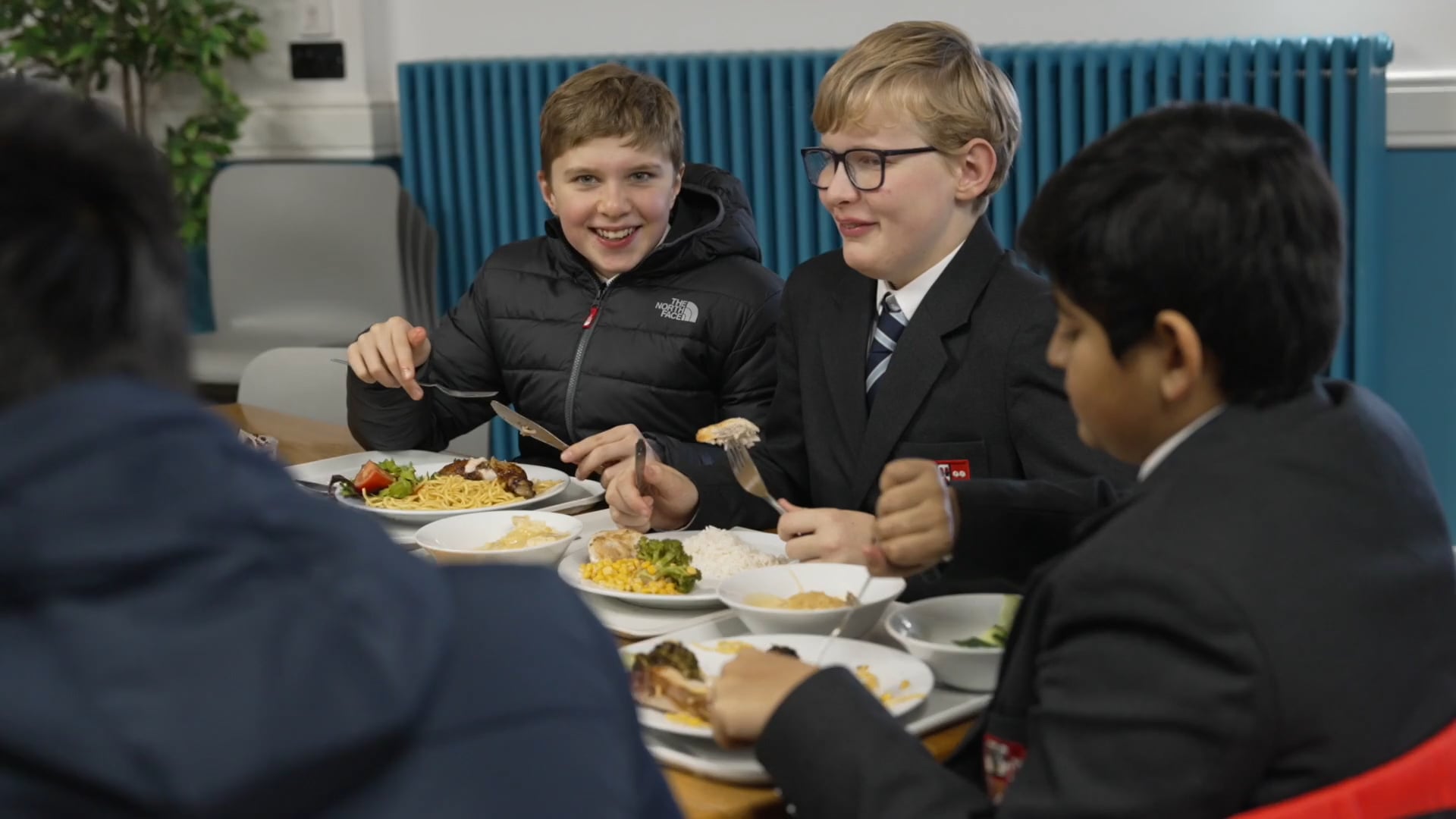 Four schoolboys in uniforms sit around a table at Brentwood School, enjoying a lively meal together. Plates of delicious Brentwood School food, including vegetables and noodles, are on the table as they share smiles and laughter.