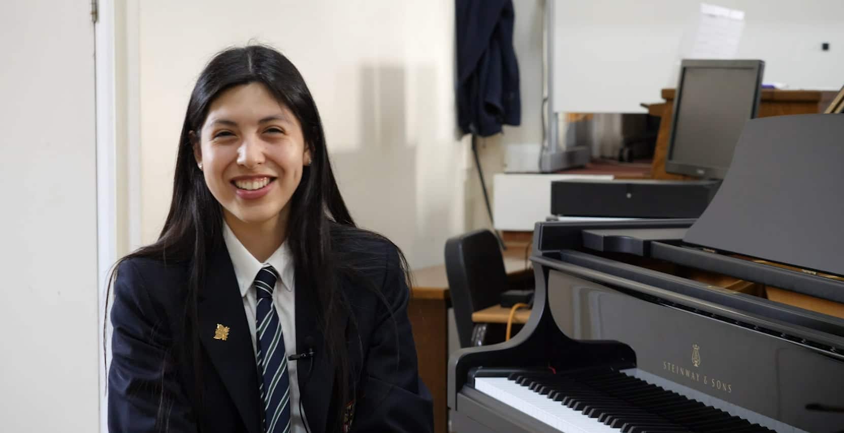 A young woman in a school uniform sits smiling beside a Steinway & Sons grand piano in a music room, talking about the Brentwood International Music Competition, with shelves, a chair, and a laptop in the background.