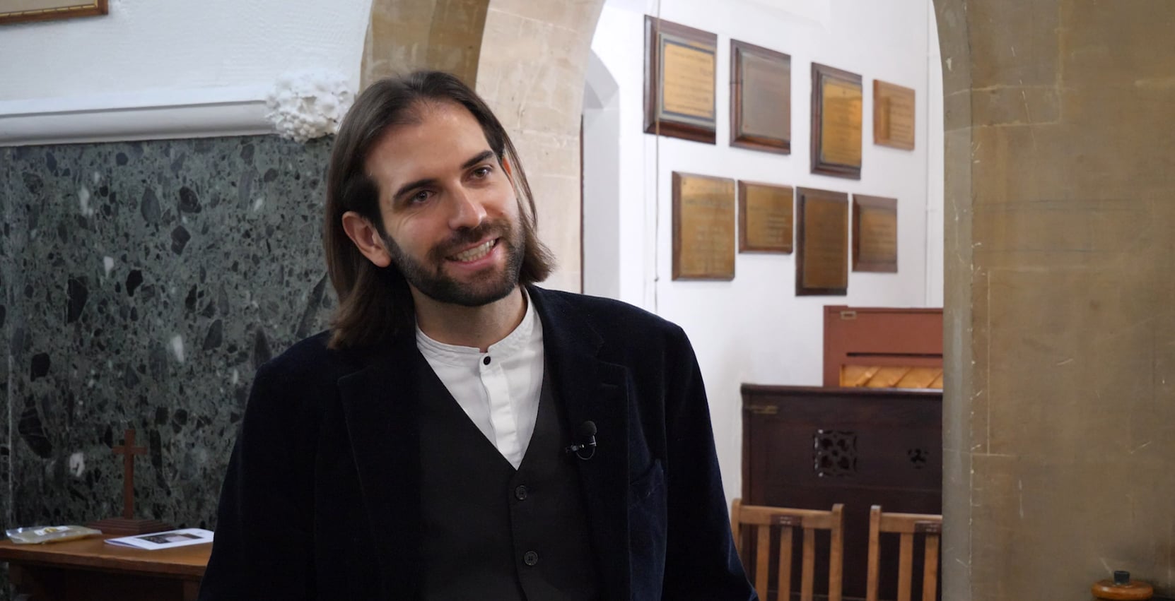 A man with shoulder-length brown hair and a beard, wearing a dark blazer and white shirt, stands smiling indoors near stone arches and wooden plaques on the wall at Brentwood School.
