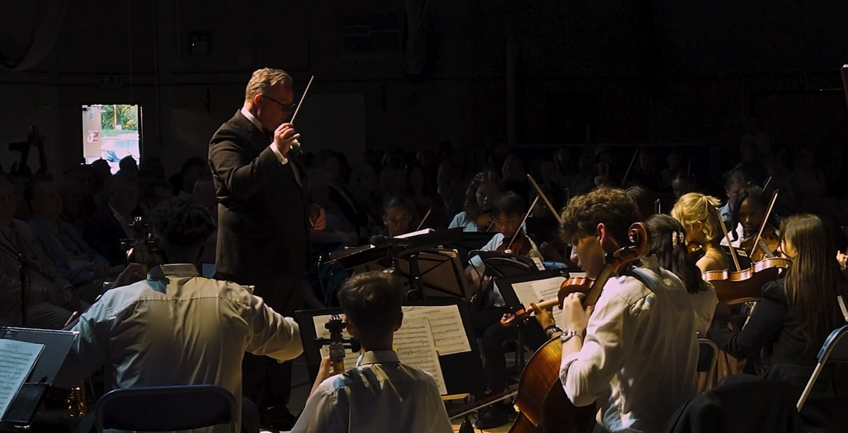 A man conducting an orchestra with students in the background at Brentwood School.