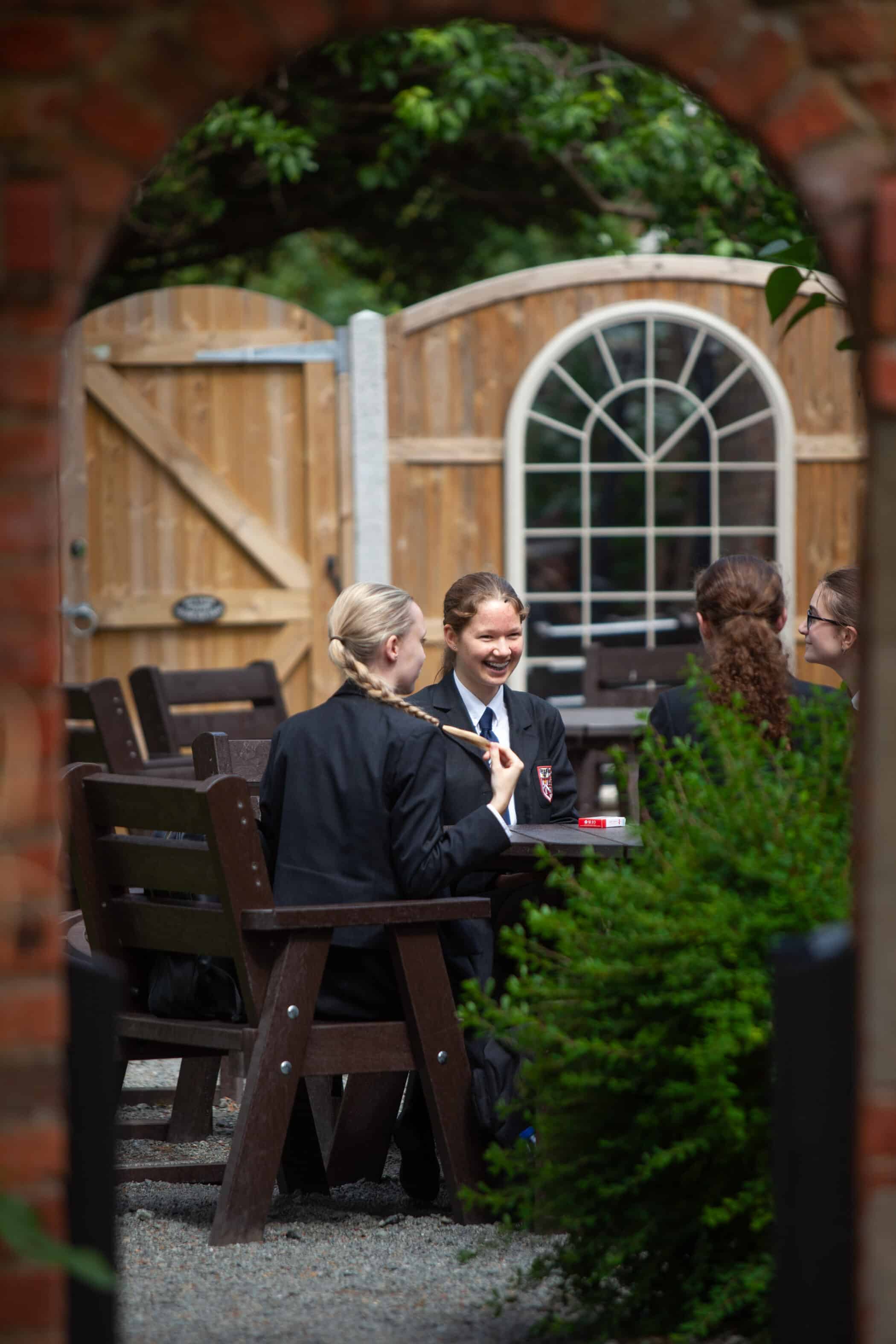 Four Brentwood School pupils in uniforms sit at a picnic table outdoors, chatting and smiling. The scene is framed by a brick archway, with a wooden fence and greenery in the background.