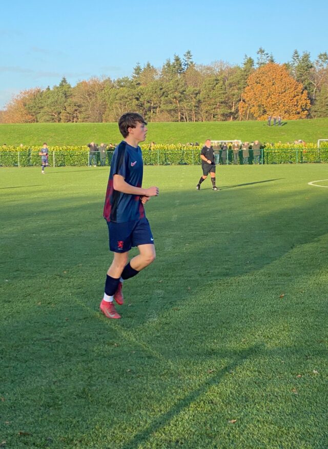 A young footballer in a navy blue kit and red boots runs on a grassy pitch during a match; trees and spectators are visible in the background under a clear sky.