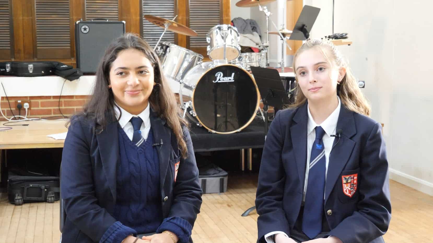 Two students in school uniforms sit side by side in a music room, with a drum kit and other instruments in the background. Both are smiling gently, discussing their recent experience at Brentwood School.