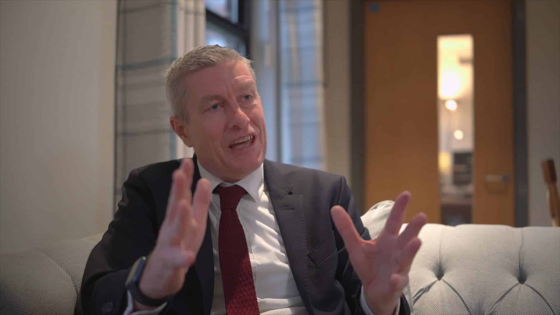 A man in a suit and red tie sits on a light-coloured sofa, gesturing as he discusses the Brentwood International Music Competition in a modern office or lounge, with a wooden door and window in the background.