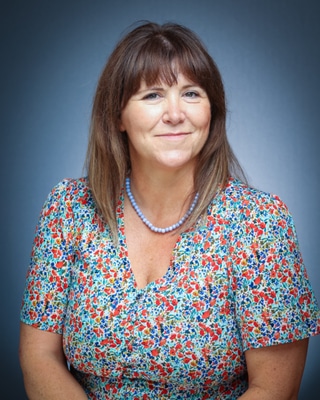 A woman with straight brown hair and fringe, wearing a pastoral floral-patterned top and a beaded necklace, smiles slightly whilst posing in front of a blue gradient background.