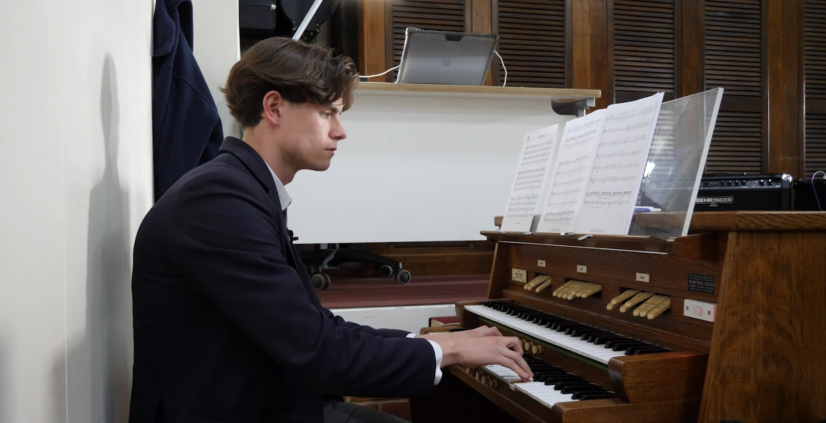 A young man in a suit at Brentwood School, plays an organ and reads sheet music propped up before him. He is seated in a small room with wooden panelling and office equipment in the background.
