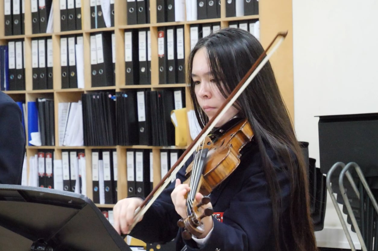 A young woman with long dark hair plays the violin indoors, focused on sheet music at Brentwood School. Behind her are shelves filled with neatly organised black and white binders.