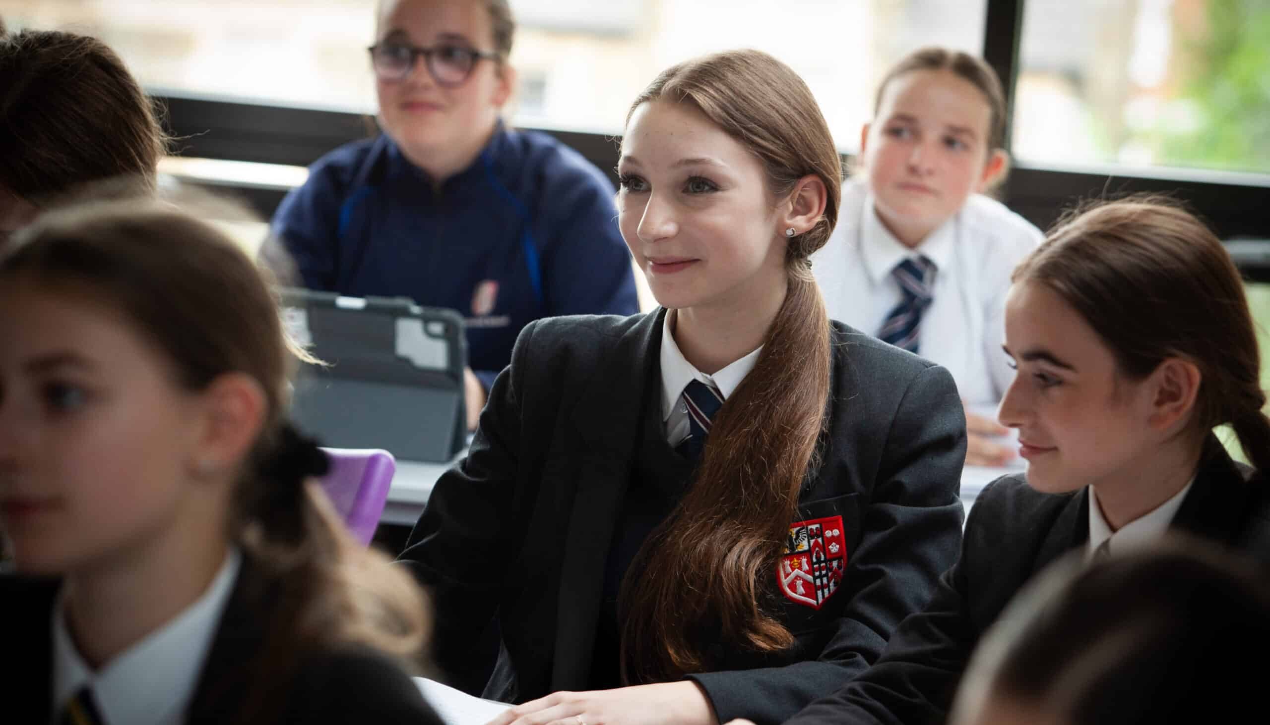 A group of students in school uniforms sit at desks in a classroom, attentively listening. One girl in the centre, smiling, sits with her hands on the desk. The academic atmosphere is clear as pupils focus, with a window visible in the background.