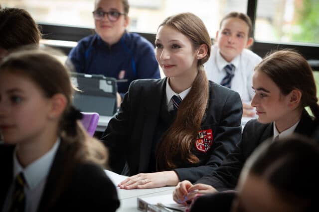A group of pupils in school uniforms sit at desks in a classroom, attentively listening to a lesson at Brentwood School. One girl in the centre is smiling, surrounded by classmates. Large windows in the background let in natural light.