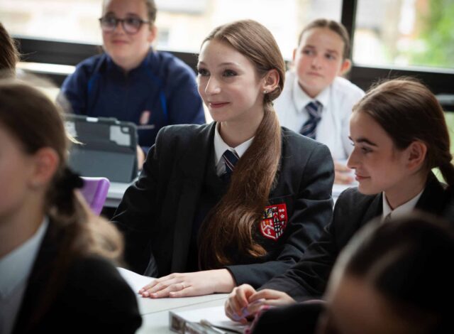 A group of pupils in school uniforms sit at desks in a classroom, attentively listening to a lesson at Brentwood School. One girl in the centre is smiling, surrounded by classmates. Large windows in the background let in natural light.