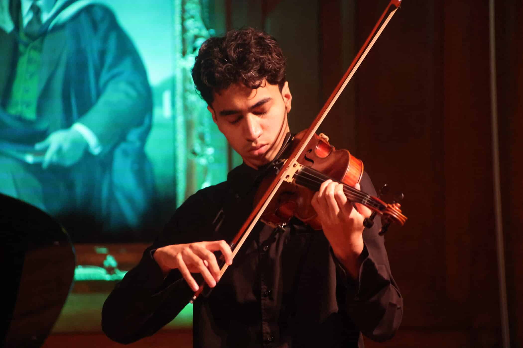 A young man in a black shirt passionately plays the violin on stage at Brentwood School, with a framed portrait in green tones visible in the background.
