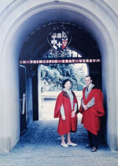 Two people in red academic robes stand under a stone archway with a crest and the Latin phrase In Principio Erat Verbum above them. Trees and a building are visible in the background.