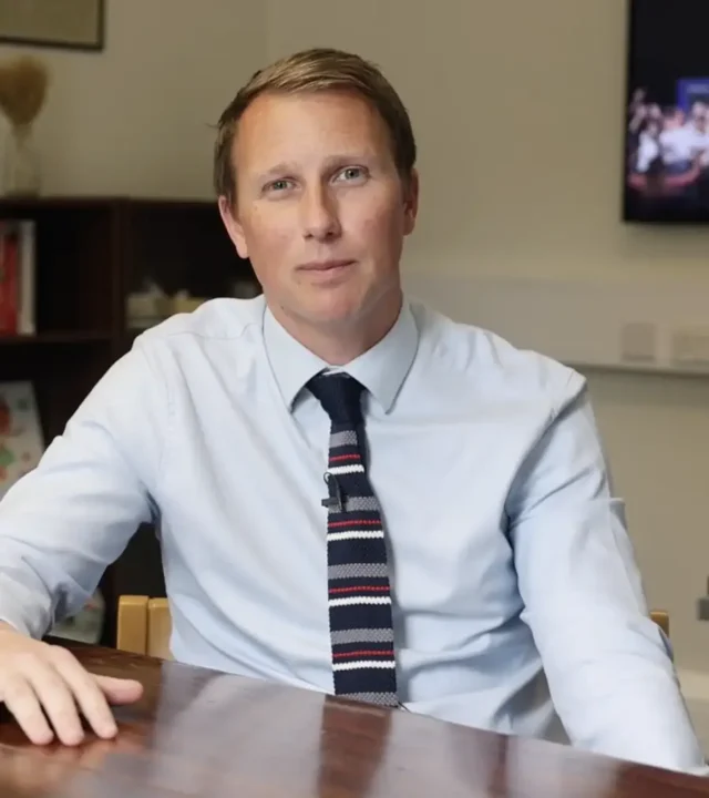 A man with short light brown hair, wearing a light blue shirt and striped tie, sits at a wooden table in a Brentwood office setting, looking directly at the camera. Shelves and a wall-mounted TV are in the background.