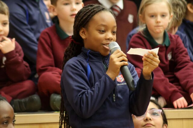 A girl holding a microphone and reading from a sheet of paper speaks to an audience, with several children seated behind her, some wearing school uniforms.