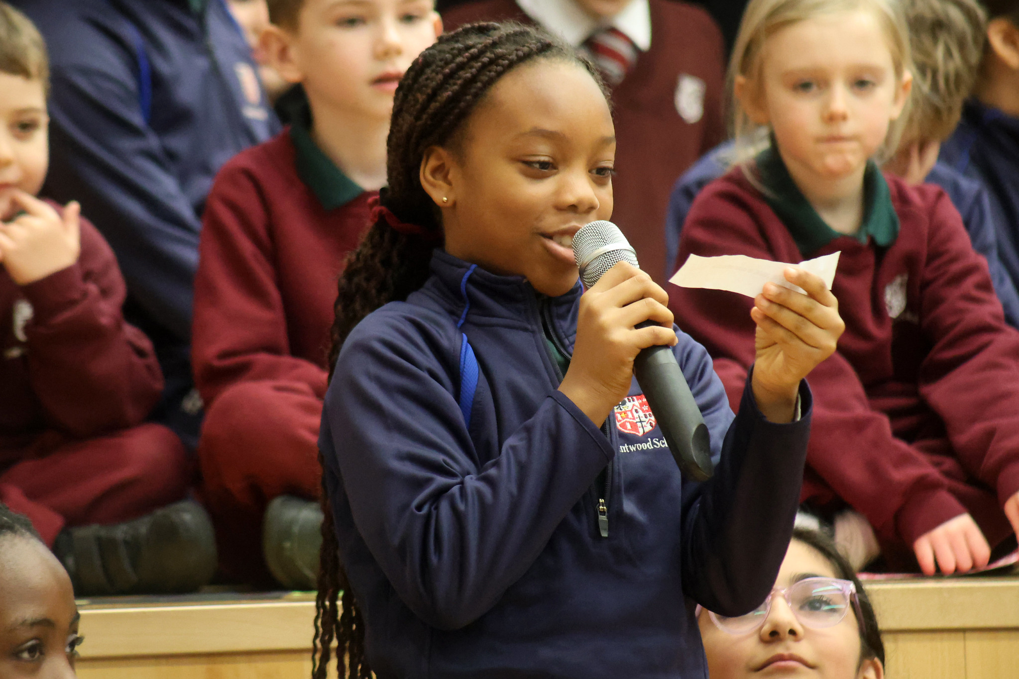 A girl holding a microphone and reading from a sheet of paper speaks to an audience, with several children seated behind her, some wearing school uniforms.