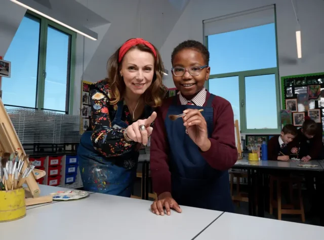 A smiling Brentwood teacher in a floral shirt and red headband stands beside a pupil holding a paintbrush in a bright art classroom, with other children working in the background.