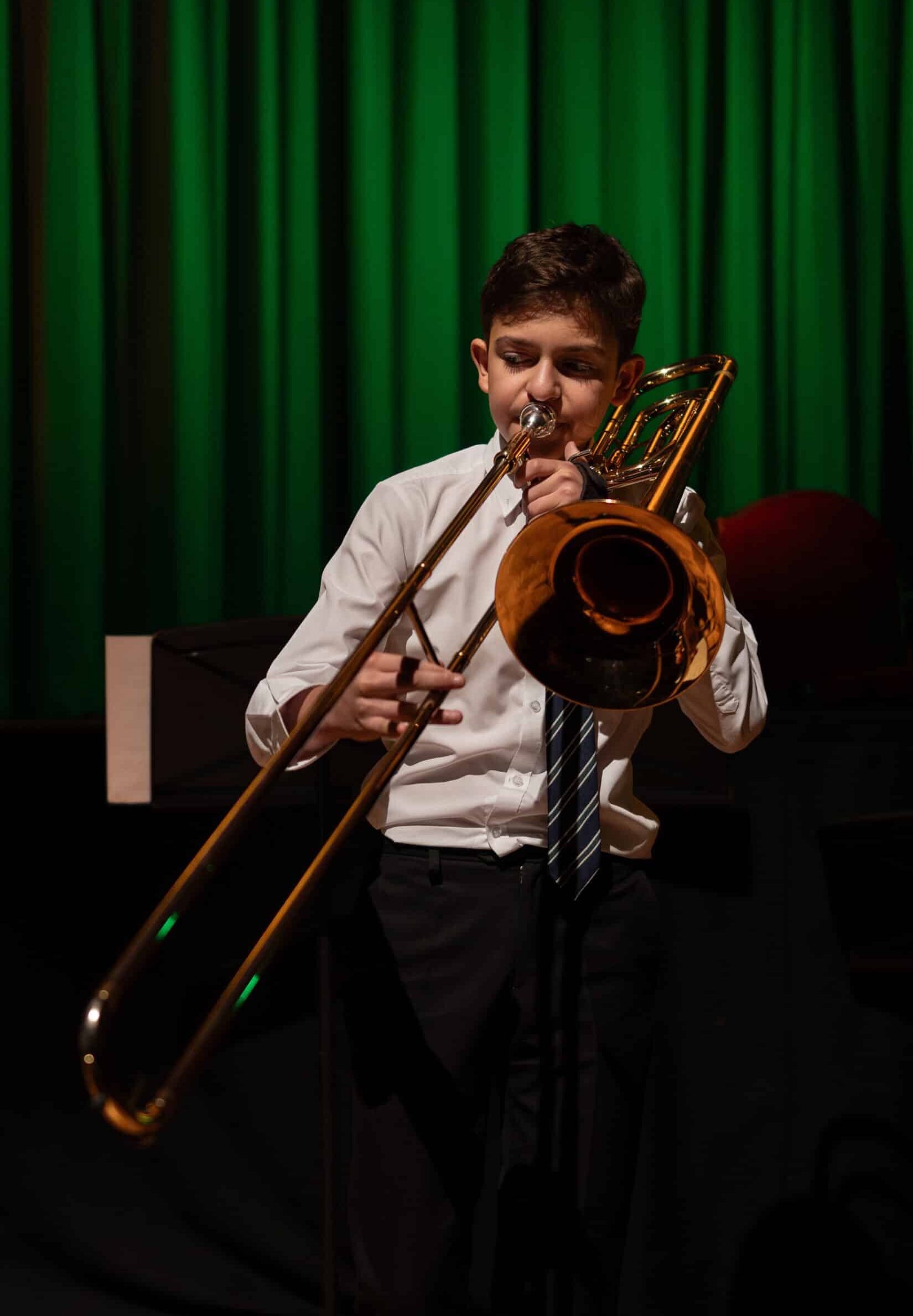 A young boy in a white shirt and tie plays a trombone on stage at Brentwood School, with green curtains behind him and dramatic spotlighting highlighting the instrument.