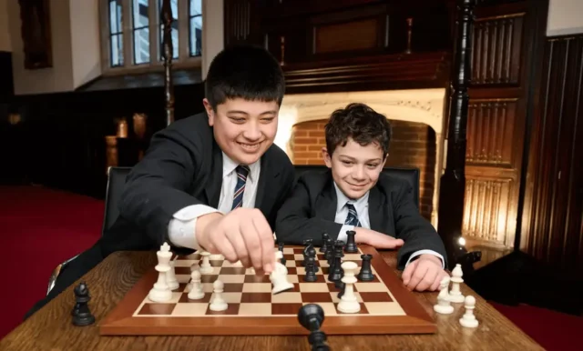 Two boys in Brentwood school uniforms play chess together at a wooden table in a warmly lit room with a fireplace. One boy smiles as he moves a chess piece, whilst the other watches intently.