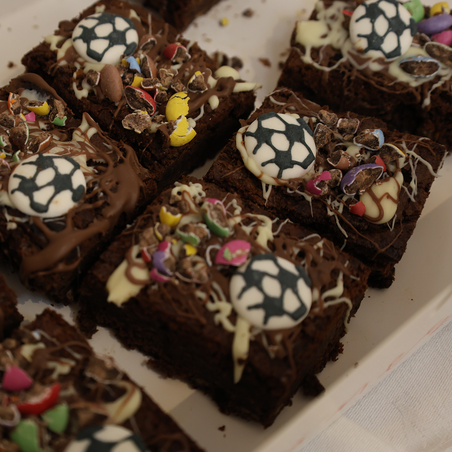 Brownie squares decorated with chocolate, white icing resembling footballs, and colourful sweet pieces, arranged on a rectangular white tray.