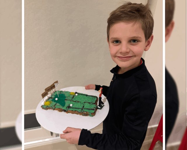 A smiling young boy holds a rectangular cake decorated to look like a golf course, complete with green icing, flags, and small white balls, whilst standing indoors.
