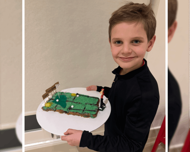 A smiling young boy holds a rectangular cake decorated to look like a golf course, complete with green icing, flags, and small white balls, whilst standing indoors.