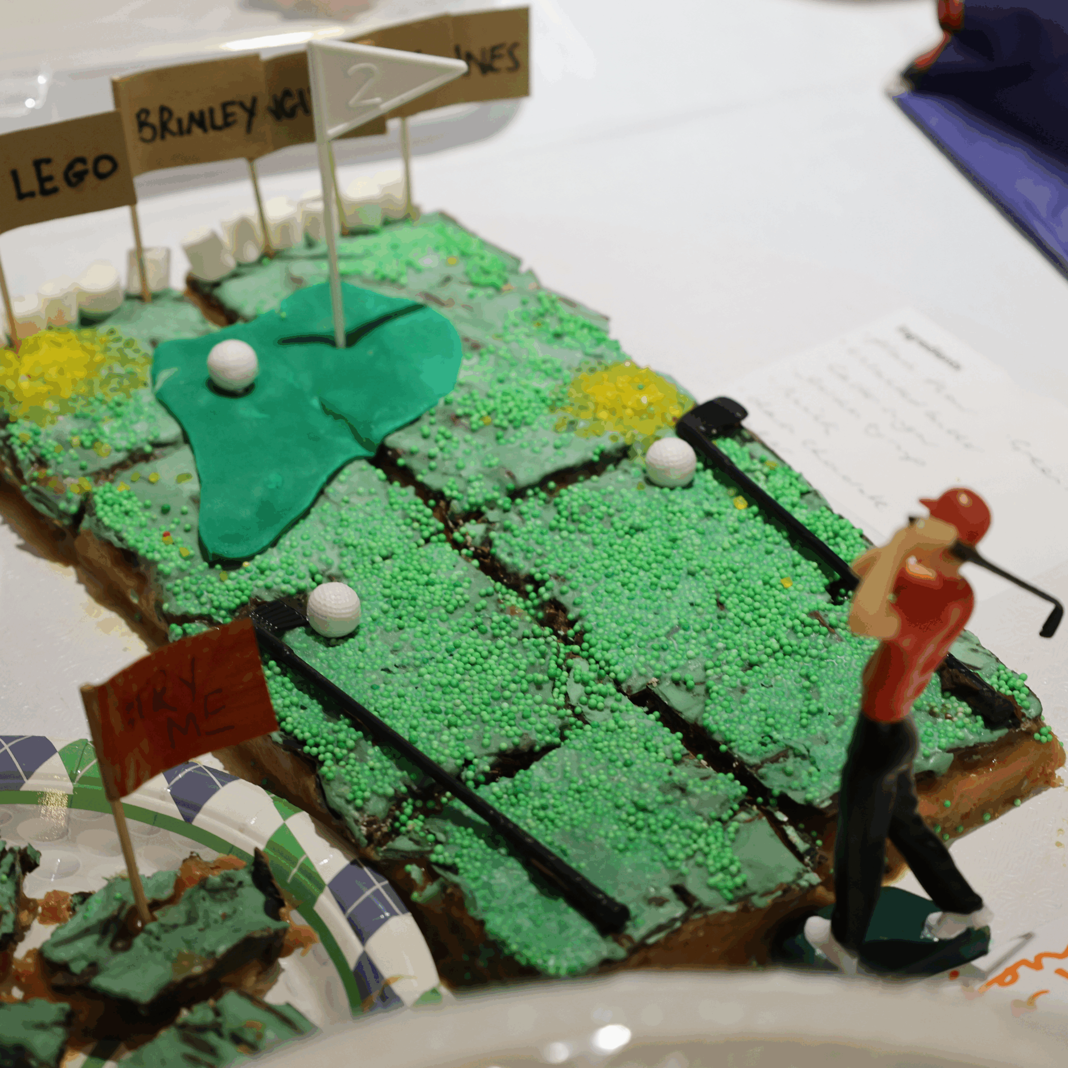 A rectangular cake decorated as a golf course with green icing, sprinkles, small flags, and golf ball sweets. A figurine of a golfer swings near the cake, and there are signs reading LEGO, BRINLEY, and 2.