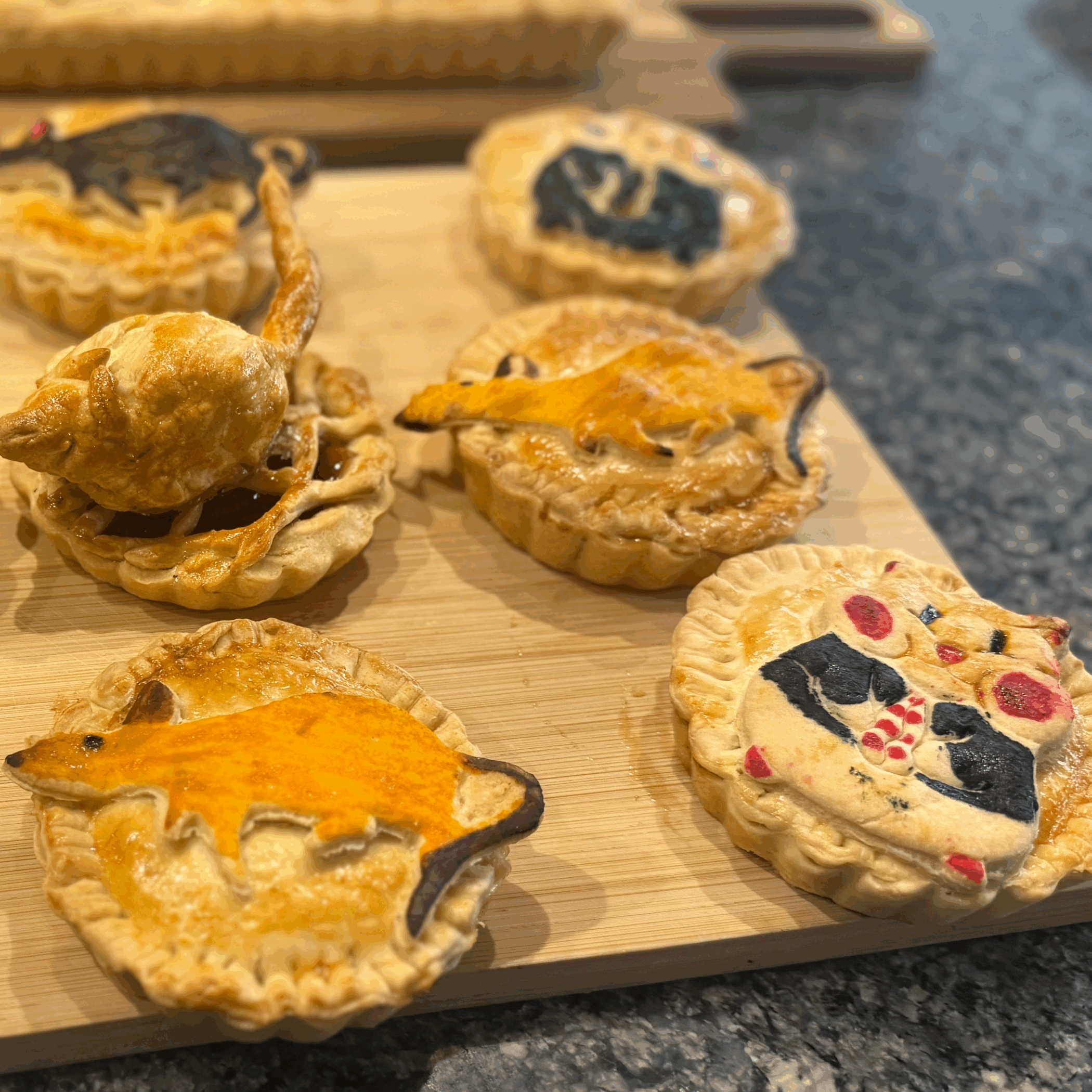 Six small pies on a wooden board, each topped with creative pastry designs of colourful animals, including a mouse, a lizard, a kangaroo, and a face with glasses and red cheeks.