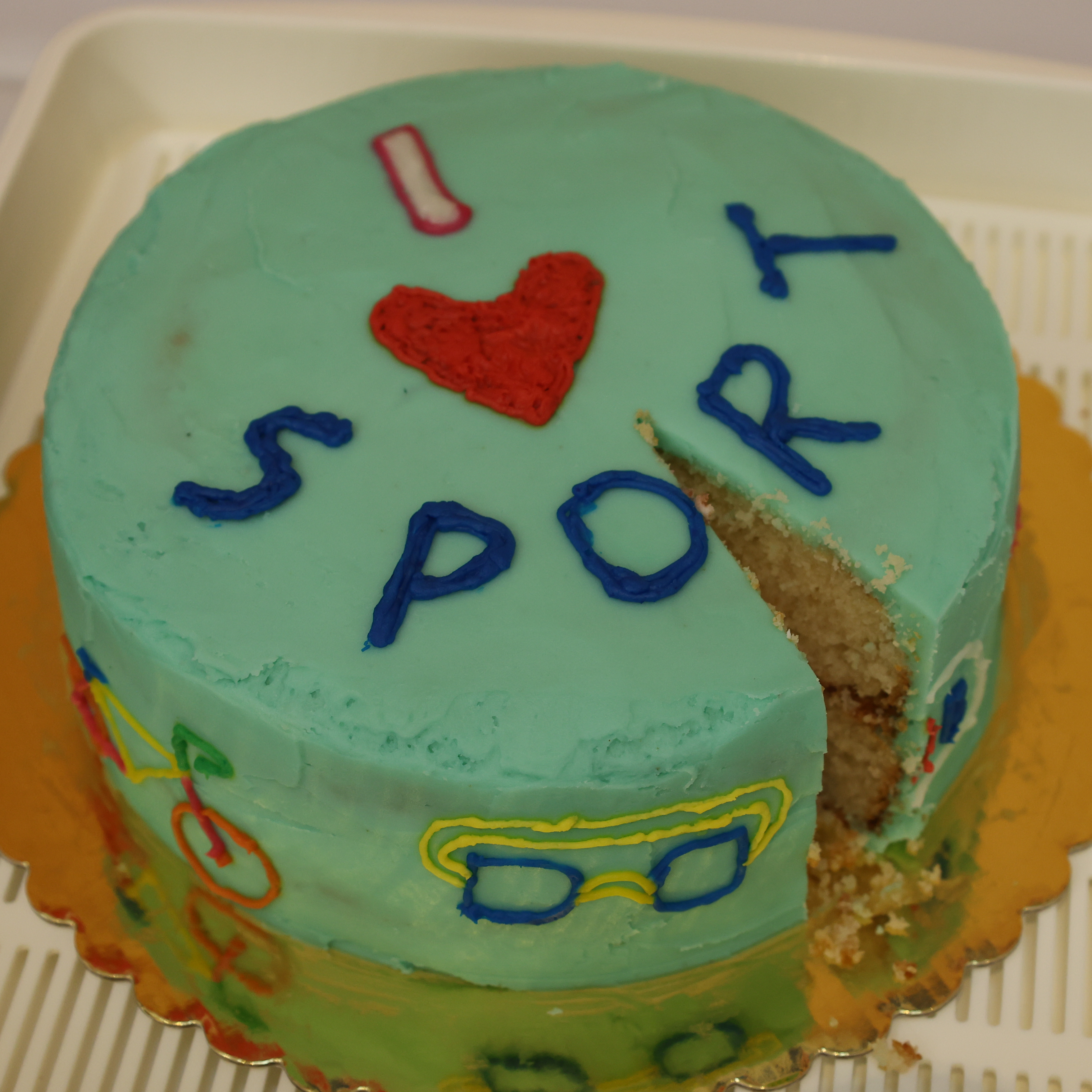 A round, light blue cake with colourful icing decorations and the text I ♥ SPORT on top. One slice has been cut from the cake, revealing the light interior. The cake sits on a gold base in a cream tray.