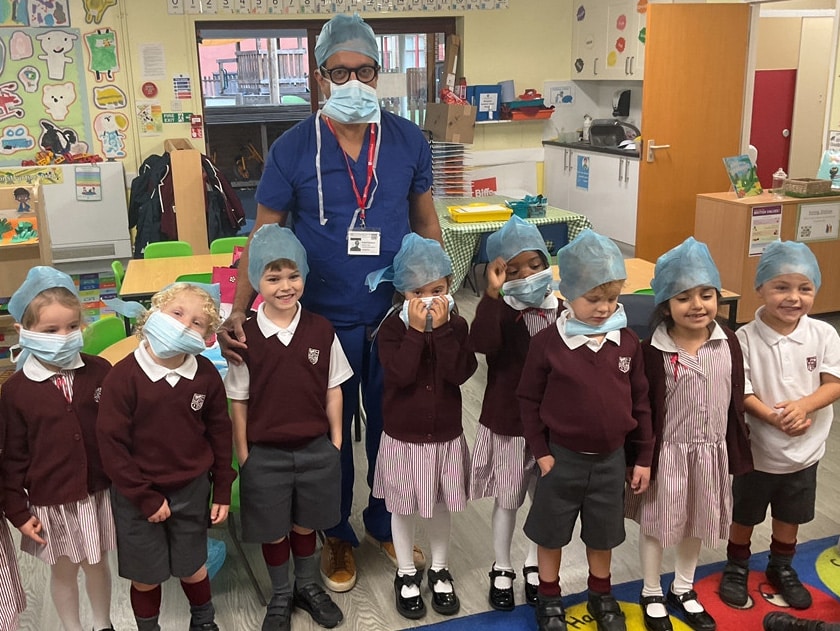 A group of young children in school uniforms stands with an adult in scrubs. Everyone is wearing blue surgical hats and face masks, smiling inside a colourful classroom decorated with drawings and educational materials.