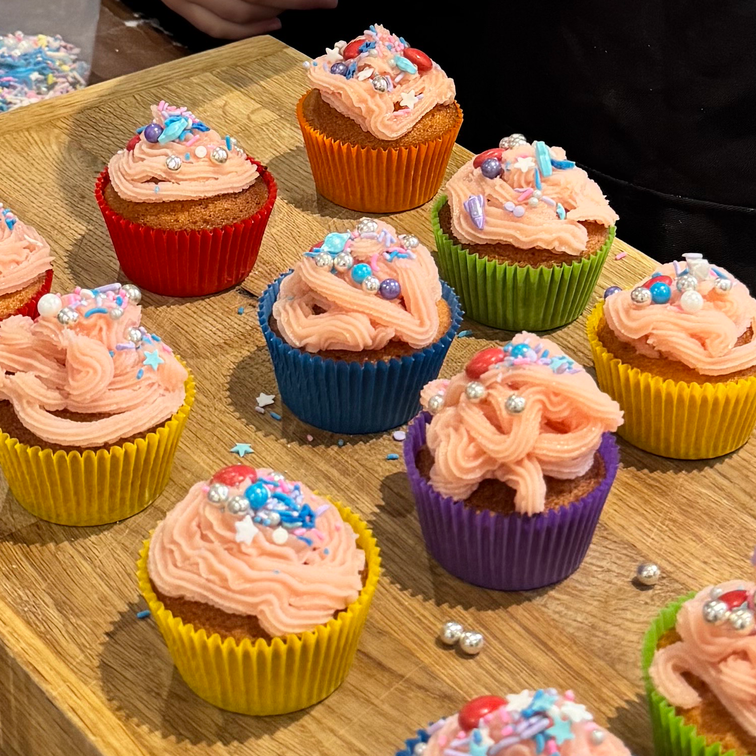 Colourful cupcakes with pink icing and assorted sprinkles are arranged on a wooden surface. The cupcakes have cases in red, blue, yellow, green, and purple. A person is partially visible in the background.
