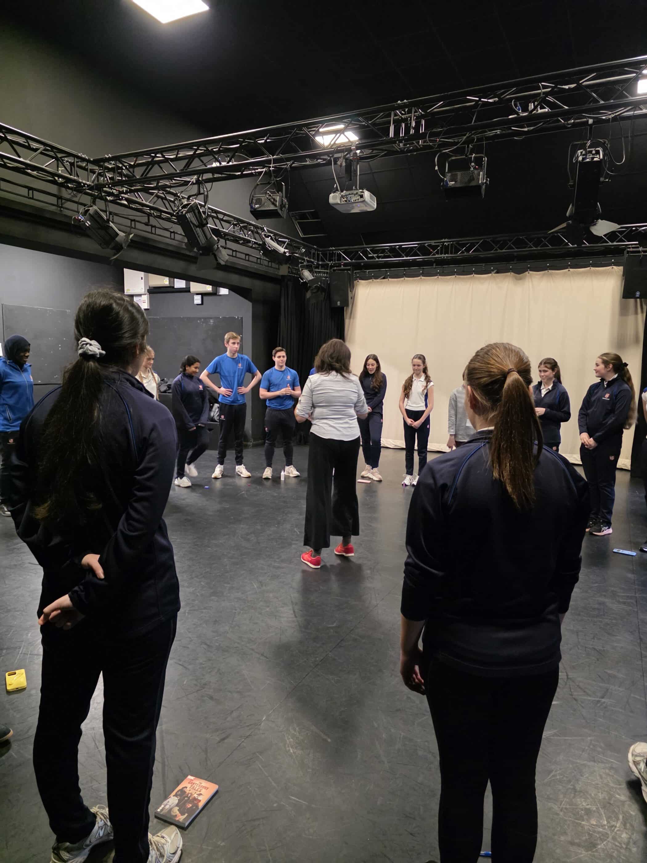 A group of people, mostly in dark sportswear, stand in a circle in a black-box theatre space, listening to an instructor in white trainers. Stage lights and equipment hang from the ceiling above them.