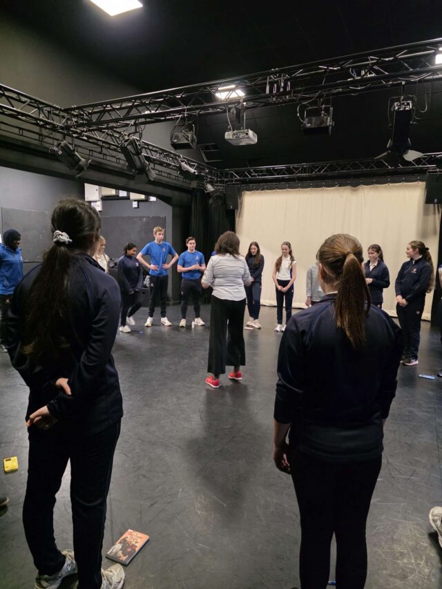 A group of people, mostly in dark sportswear, stand in a circle in a black-box theatre space, listening to an instructor in white trainers. Stage lights and equipment hang from the ceiling above them.