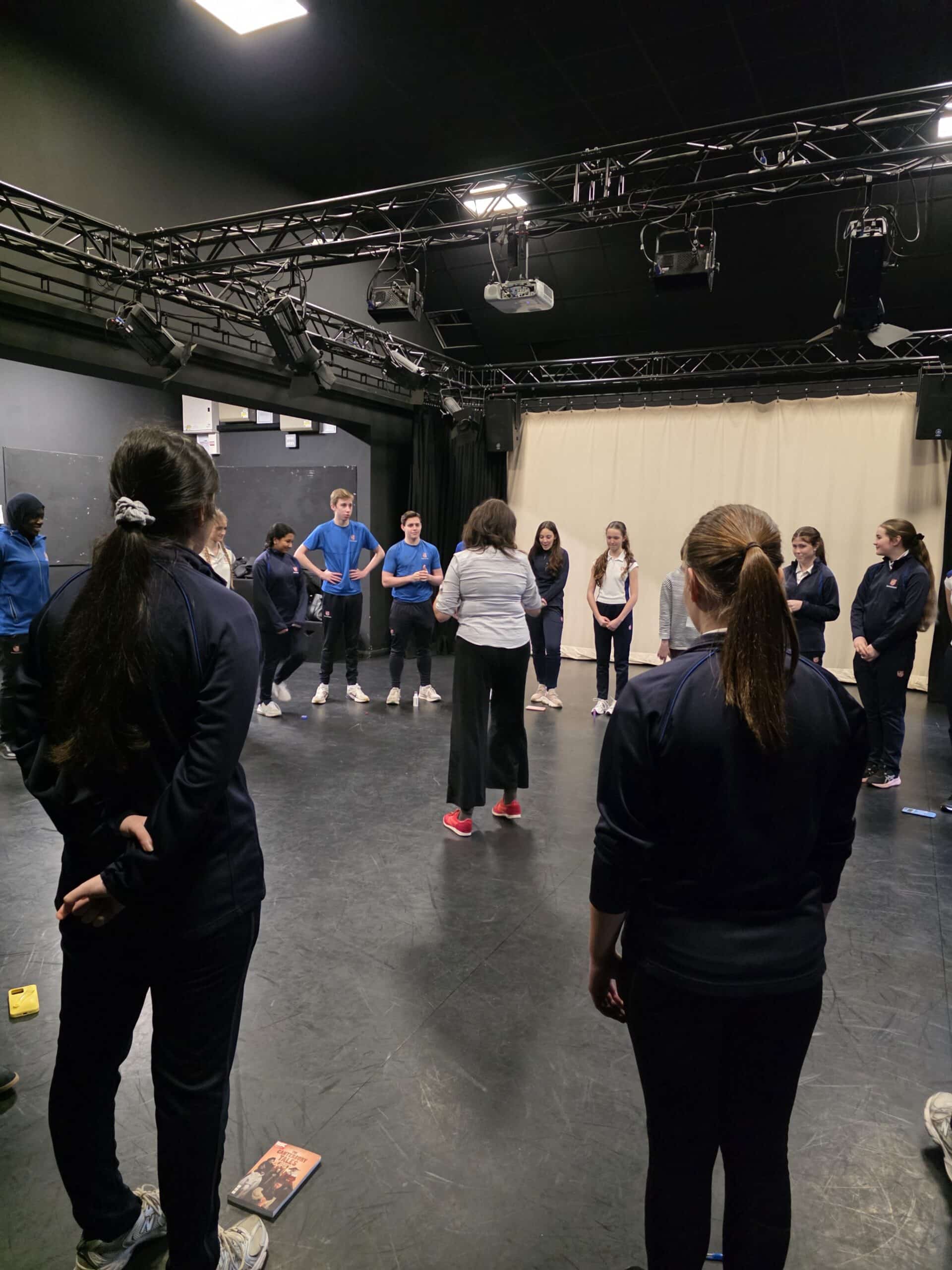 A group of people, mostly in dark sportswear, stand in a circle in a black-box theatre space, listening to an instructor in white trainers. Stage lights and equipment hang from the ceiling above them.