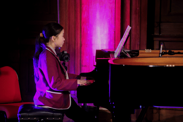 A young girl in a maroon jacket plays a grand piano on stage, reading sheet music. The scene is lit with purple and pink lighting, creating a vibrant atmosphere. A camera hangs round her neck.