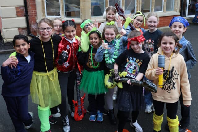 A group of children dressed in colourful 1980s-themed costumes and accessories, posing and smiling outdoors; some hold toy guitars and a toy microphone, looking happy and excited.