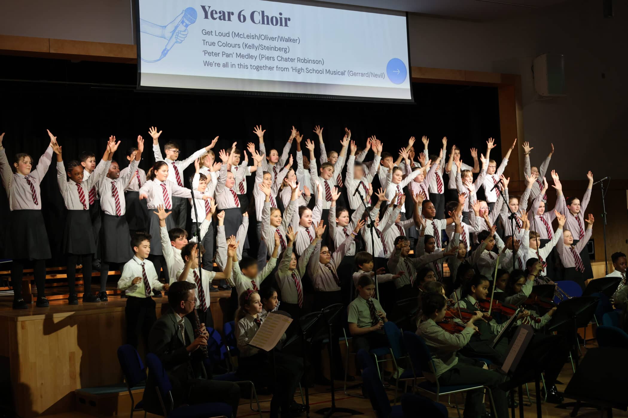 A large group of schoolchildren in uniform stand on stage singing with arms raised, whilst a seated orchestra plays in front; a screen above reads “Year 6 Choir” with a song list.