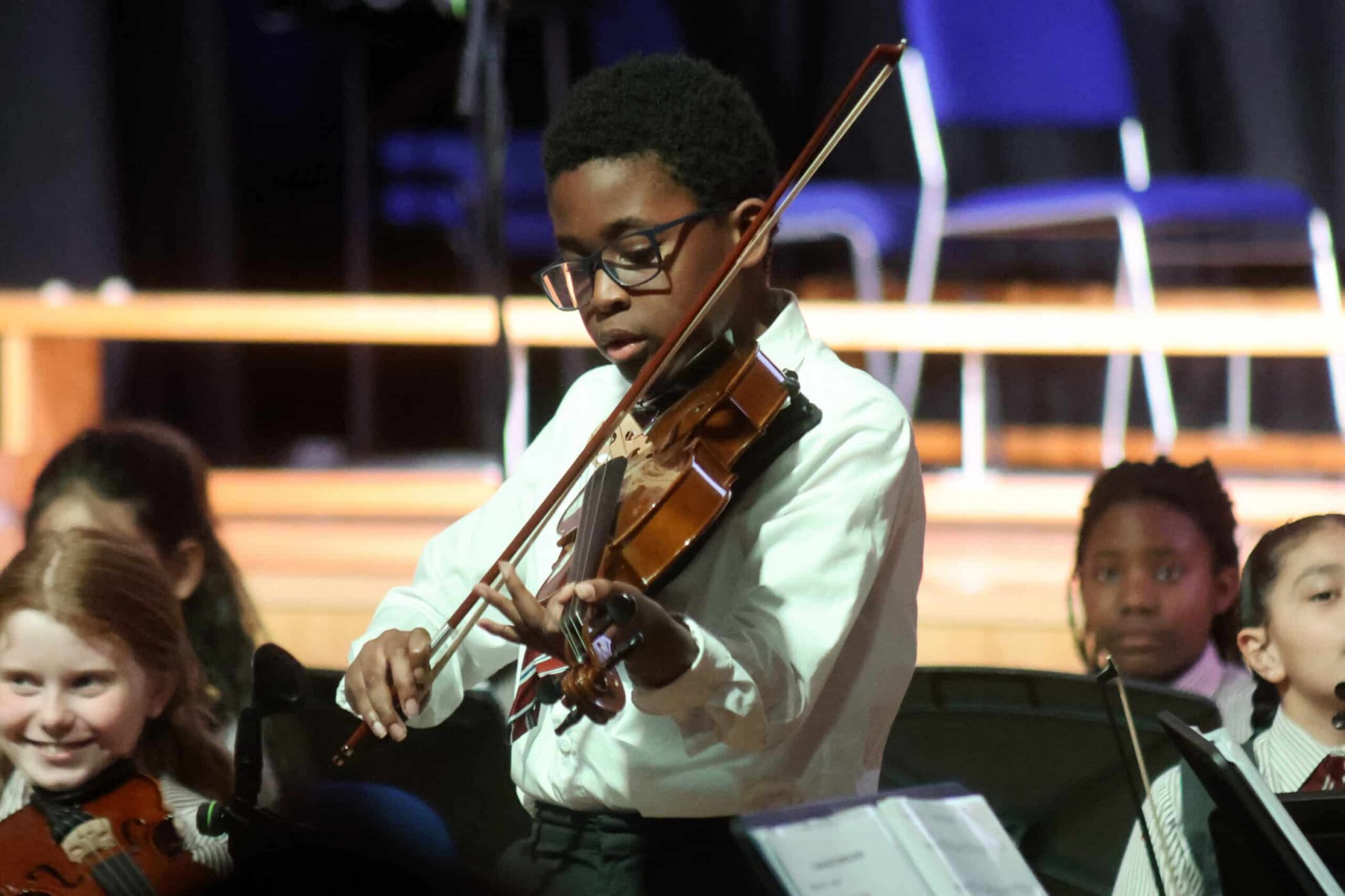 A young boy wearing glasses and a white shirt plays the violin during a concert, with other children and empty chairs visible in the background.