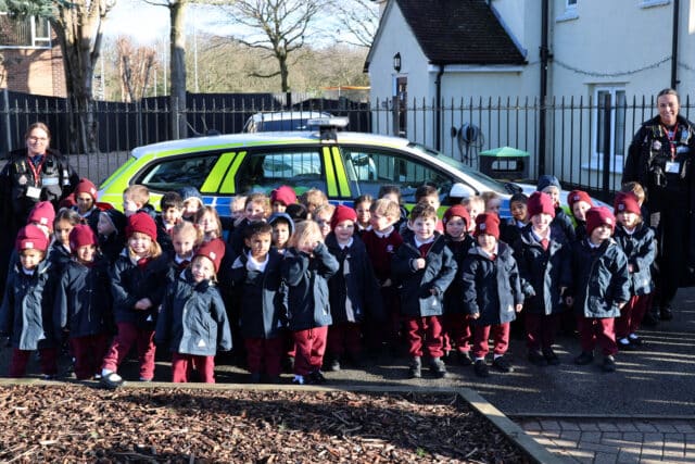 A large group of young children in matching uniforms and maroon hats stand outside in front of a police car, accompanied by three adults in black uniforms, near a white building and a black metal fence.